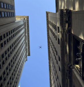 A drone piloted by AeroSpect soars between two skyscrapers in NYC, performing a building facade inspection